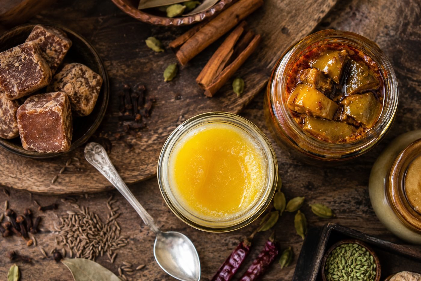 Indian pantry staples on a wooden surface: a jar of ghee, blocks of jaggery, whole spices and a jar of mango achar.