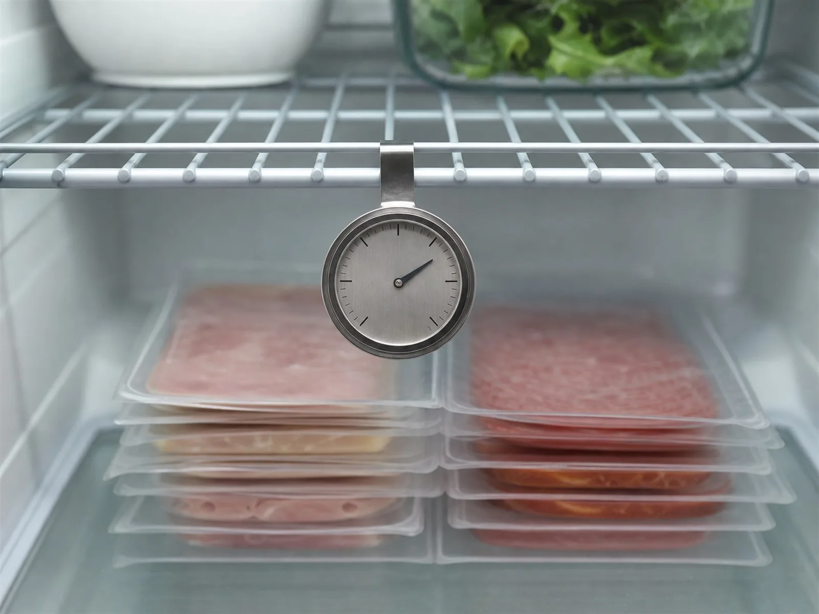 Refrigerator thermometer reading 38&deg;F beside stacked deli meat packages stored on a lower shelf, away from the door