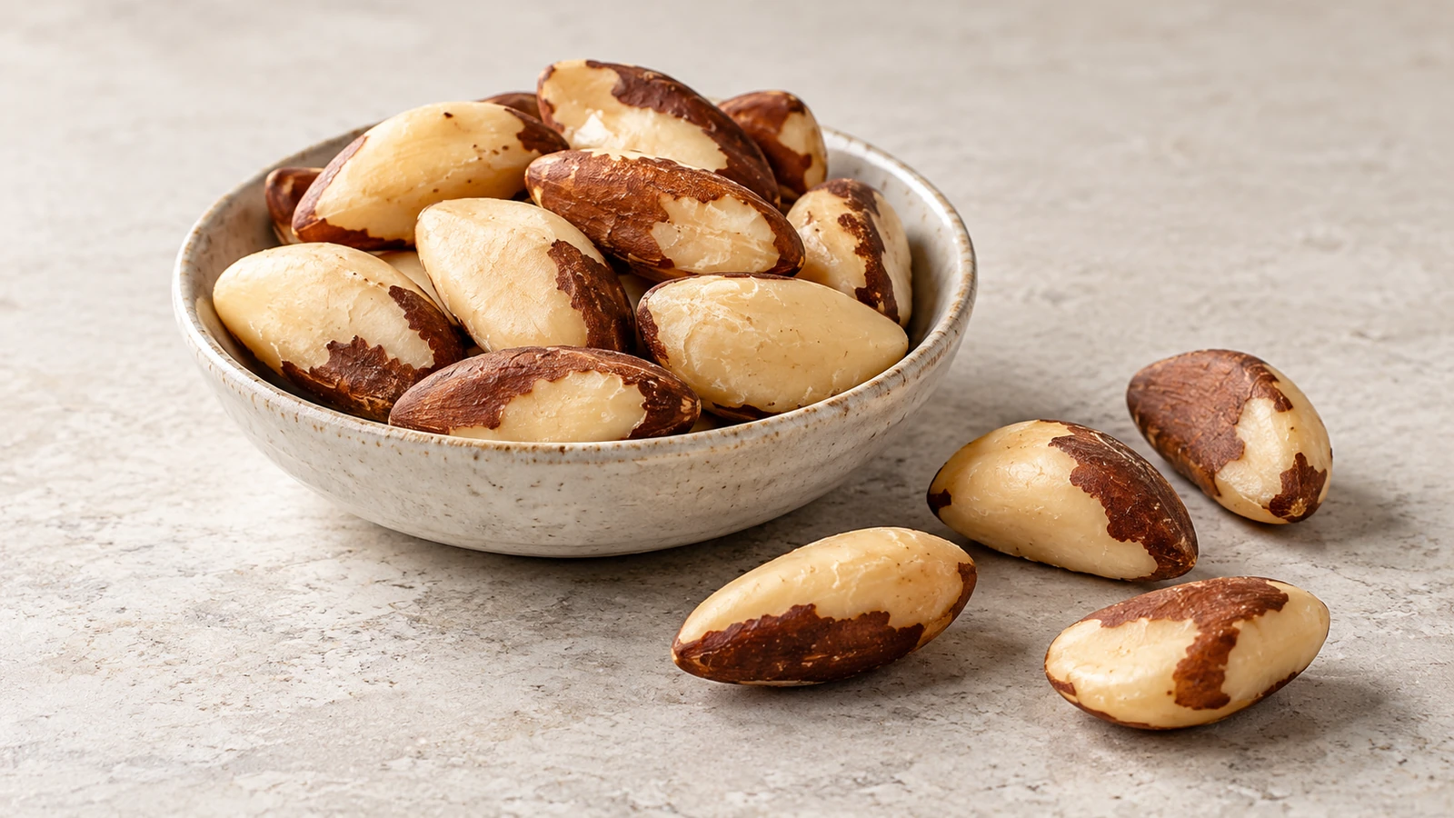 Shelled Brazil nuts with cream interiors and brown skin in a ceramic bowl and on a stone surface.