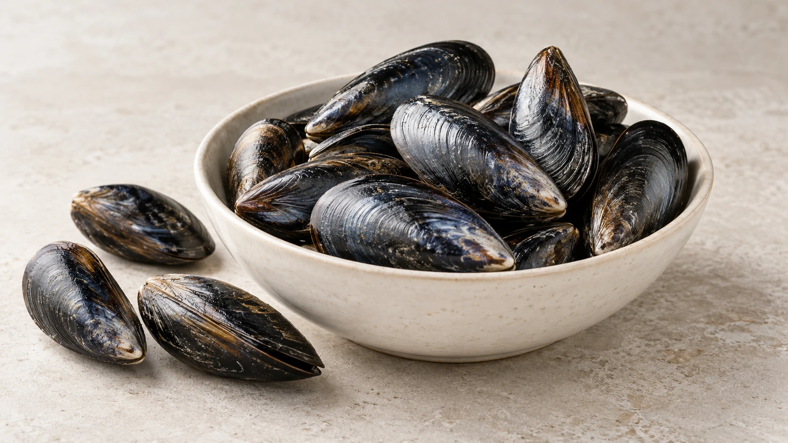 Blue-black mussel shells in a ceramic bowl with three shells beside it.