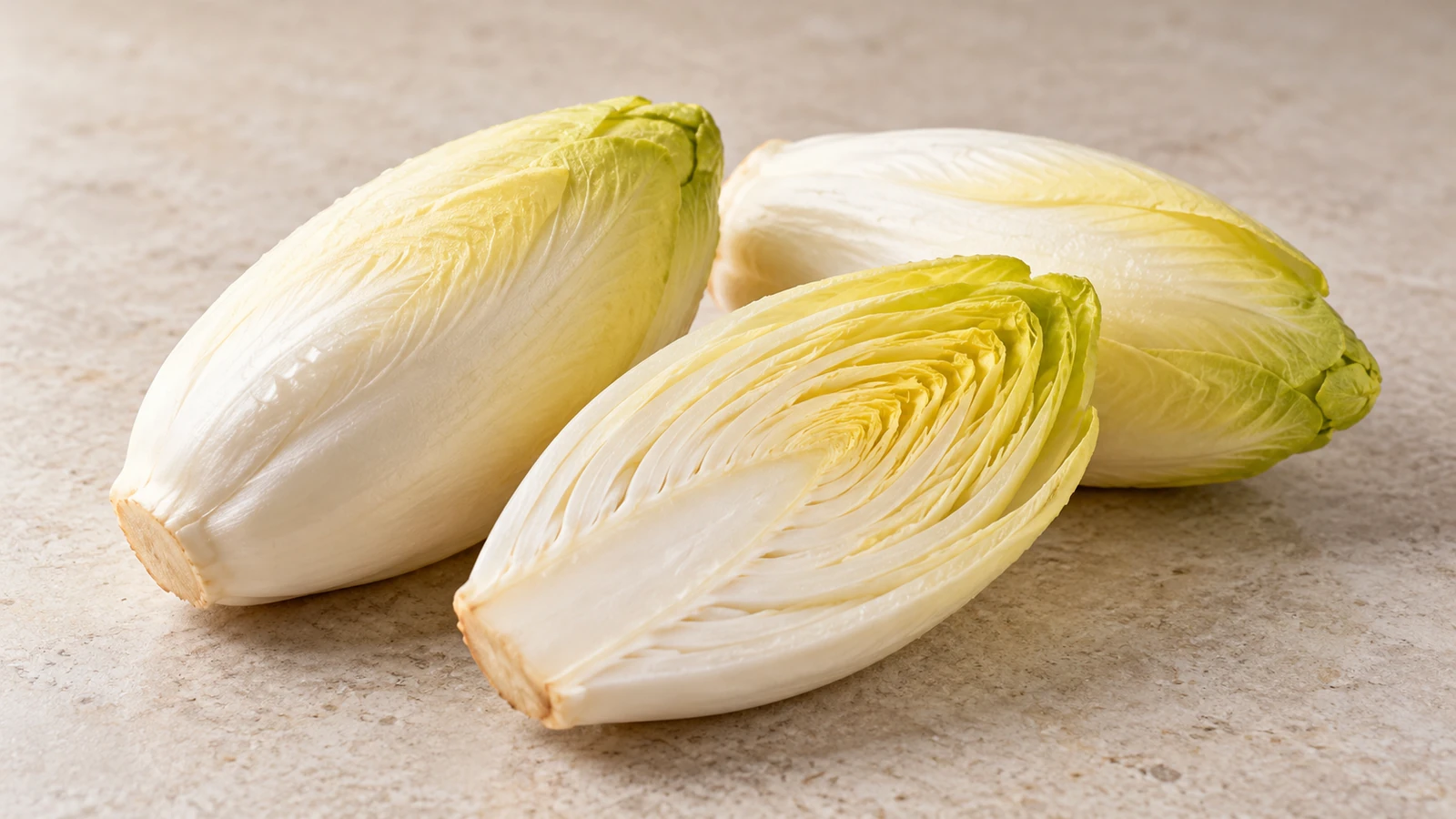 Two whole Belgian endive heads beside a lengthwise half showing tightly layered pale leaves.